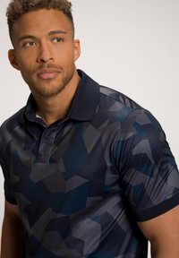 Polo à manches courtes en tissu lisse de couleur marine, présentant un motif géométrique dans des nuances variées de bleu et de gris ; comprend un col et une patte de boutonnage.
