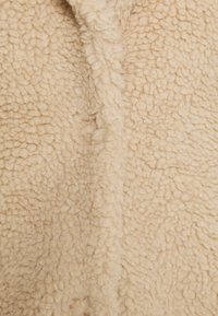 Beige fleece-stof med en blød, tekstureret overflade, der har et krøllet mønster og et varmt, hyggeligt udseende. Velegnet til ydertøj.