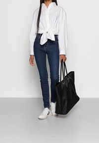 Vit skjorta knuten i midjan, mörkblå smala jeans och svart axelväska. Tillbehör inkluderar vita sneakers med svarta detaljer.