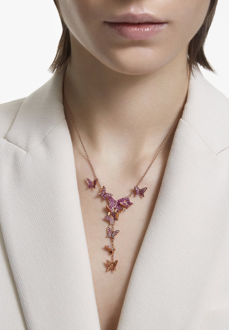 Gros plan d'une personne portant un blazer blanc et un collier en or rose avec des breloques papillons roses et en or rose suspendues en forme de Y.