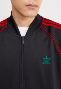 Svart zip-up jacka med röda axelränder och grön Adidas-logotyp. Slät tyg och ribbad krage som framhäver den atletiska designen.