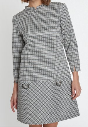 Frau trägt ein graues Kleid mit Hahnentrittmuster, langärmlig, mit kleinen vorderen Taschen, die silberne Verschlussdetails aufweisen.