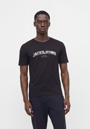 Mand med kort hår iført en sort Jack & Jones t-shirt og mørke bukser, stående mod en ensfarvet hvid baggrund.