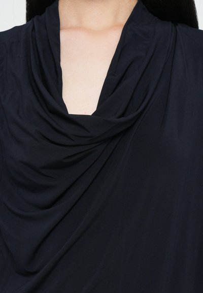 Gros plan d’une personne portant un haut noir à col bénitier drapé, montrant le décolleté et la partie supérieure de la poitrine.