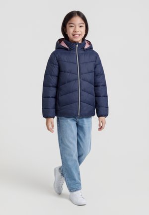 Enfant souriant et marchant portant une veste matelassée bleu marine avec capuche, un jean bleu clair et des baskets blanches sur un fond uni clair.