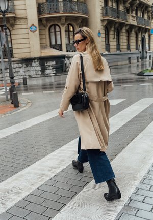 Beżowy trencz z paskiem w talii, w połączeniu z dżinsowymi spodniami w stylu dzwonów i czarnymi botkami za kostkę. Dodatki obejmują małą czarną torbę i okulary przeciwsłoneczne.