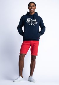 Marine hoodie met witte print, rode shorts en witte sneakers. De hoodie heeft een trekkoord en een zak aan de voorkant. Glad stoftextuur.