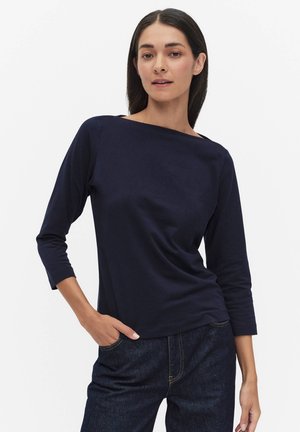 Kvinde iført en mørkeblå langærmet top og mørke jeans, poserer med den ene hånd i lommen foran en ensfarvet hvid baggrund.