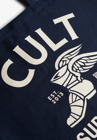 Granatowa torba typu tote z płótna, z wyraźnym białym napisem "CULT" oraz ilustracją skrzydlatej boot. Wytrzymały materiał, wyrazisty design graficzny.