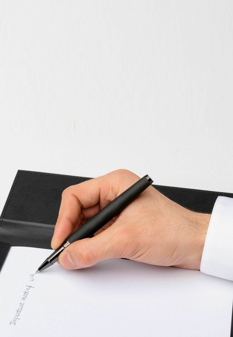 Čierne pero držené v ruke, píše na bielej papieri v notes. Obal notesu je čierna koža s hladkým povrchom.