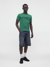 Άνδρας που φοράει πράσινο t-shirt Dsquared2 Milano, τζιν σορτς μήκους έως τα γόνατα με αλυσίδα, λευκές κάλτσες και μαύρα αθλητικά, στέκεται σε απλό φόντο.
