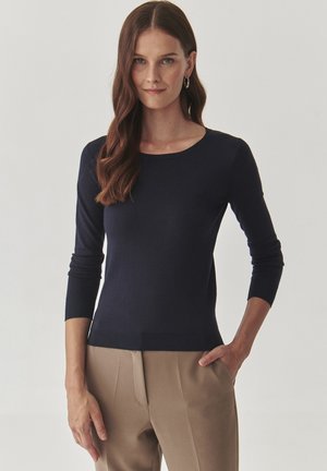 Donkerblauwe gebreide trui met lange mouwen, een ronde halslijn en geribde boorden, gecombineerd met beige broek. Soepele textuur, aansluitend ontwerp.