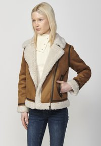 Giacca in shearling marrone con un ampio colletto e polsini in crema, dotata di una chiusura a zip e bordature scure in contrasto lungo i bordi.