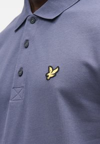 Polo em tecido azul escuro com acabamento texturizado, apresentando três botões e um pequeno logótipo bordado de um pássaro amarelo no peito.