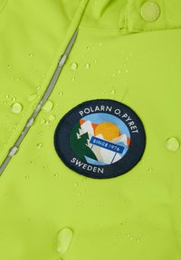 Vihreä vedenpitävä takki, jossa on pyöreä merkki, jossa näkyy vuoria, aurinko ja vettä, ja joka on merkitty "POLARN O. PYRET SWEDEN", pinnalla vesipisaroita.
