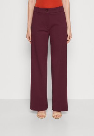 Pantalons larges bordeaux en tissu lisse, avec passants de ceinture et fermeture à bouton, associés à des sandales ouvertes beiges claires.