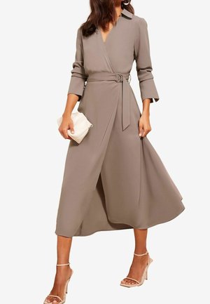 Kobieta w taupe sukience kopertowej z rękawami do łokcia, z wiązanym w pasie, trzyma białą kopertówkę i ma na sobie cieliste sandały na wysokim obcasie.