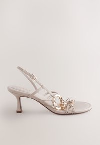 Escarpin à talon beige avec des brides fines, un accent décoratif en chaîne dorée, et une surface de chaussure lisse légèrement texturée.