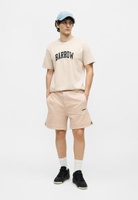 Νέος άνδρας φορώντας μπεζ t-shirt και σορτς Barrow, μπλε καπέλο, λευκές κάλτσες και μαύρα αθλητικά παπούτσια, στεκόμενος με τα χέρια στις τσέπες.