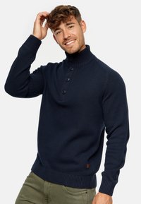 Marineblå pullovertrøje med høj krave, fire knapper, ribstrikkede ærmer og kanten. Lavet af et struktureret strikstof.