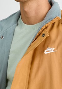 Färgblockerad jacka med ett orange yttre lager och en blå fleecekrage, parat med en ljusgrön t-shirt. Har en vit Nike-logga.