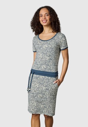 Kvinde med krøllet hår iført en kortærmet, knælang kjole med blå og hvide blomsterprint og blå kant, stående med den ene hånd i lommen.