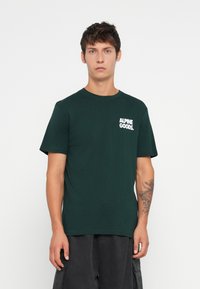 Ung mann med kort brunt hår iført en mørkegrønn "ALPINE GOODS" t-skjorte og mørke bukser, stående mot en hvit bakgrunn.