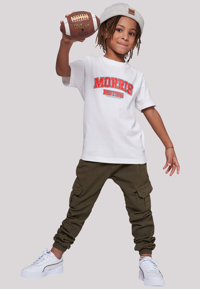 White short-sleeve t-shirt with "MORRIS MOTORS" print, olive cargo pants, and white sneakers. Model holds a brown football.
