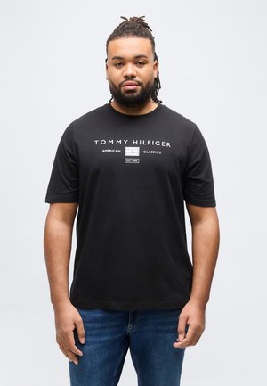 Mann mit geflochtenem Haar und Bart, der ein schwarzes Tommy Hilfiger T-Shirt und blaue Jeans vor einem schlichten hellen Hintergrund trägt.