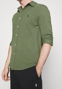 Mężczyzna w długim rękawie zielonej koszuli zapinanej na guziki z podwiniętymi rękawami oraz czarnych spodniach, z małym haftowanym logo na obu ubraniach.