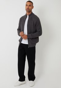 Chaqueta gris de punto con cremallera y un patrón texturizado, con puños y cinturilla de canalé, llevada sobre una camiseta blanca y combinada con pantalones negros.