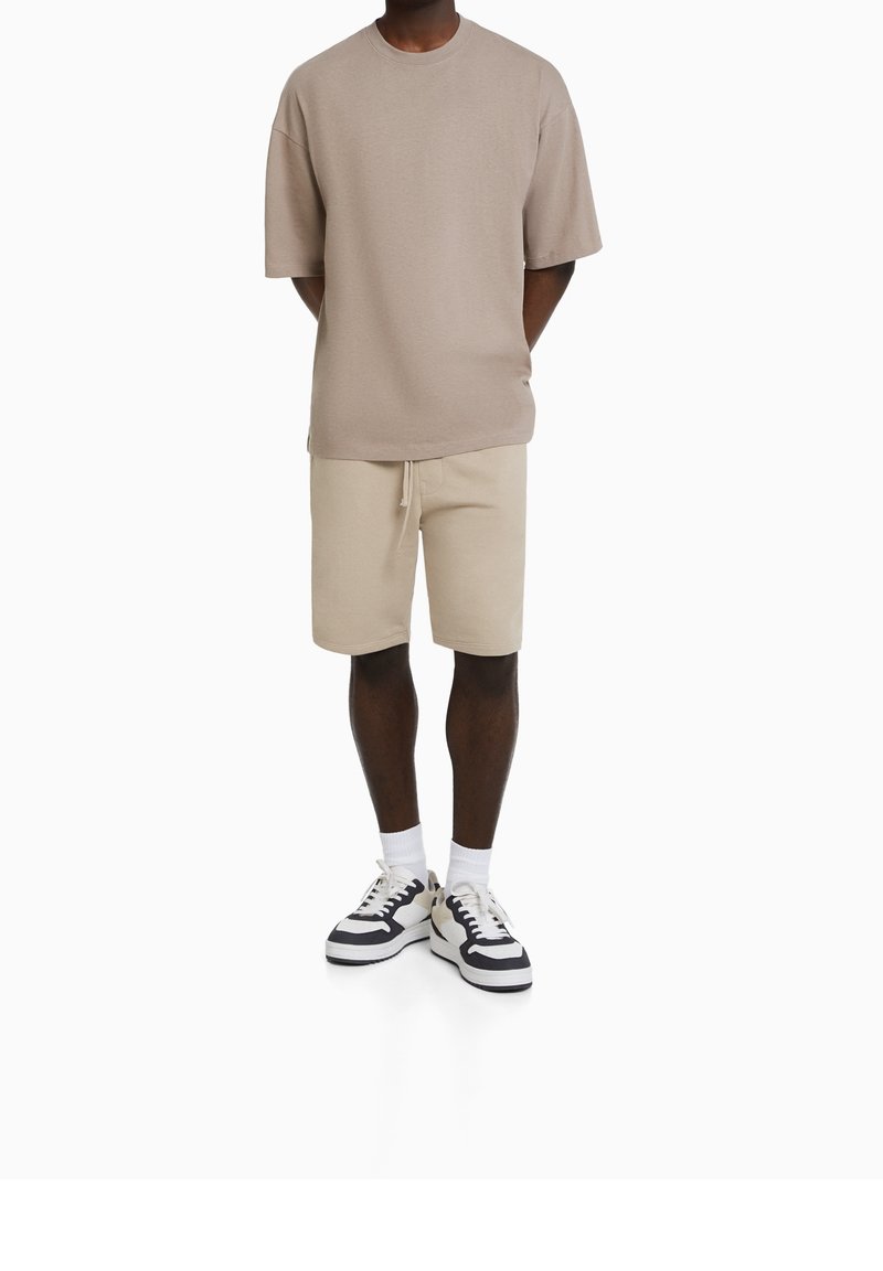 Homme portant une chemise beige oversize à manches courtes, un short assorti à cordon, des chaussettes blanches et des baskets noir et blanc, debout les mains dans le dos.