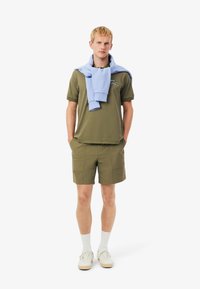 Polo verde oliva, maglione azzurro chiaro annodato intorno alle spalle, pantaloni corti oliva, calzini bianchi, sneaker beige con un design semplice.