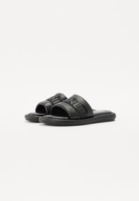 Sorte slidesandaler med bred stropp som har preget merke, glatt lærtekstur og flat gummisåle.