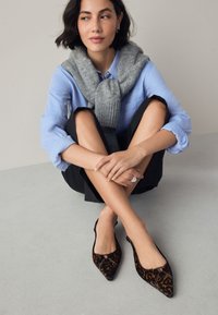 Ballerines à bout pointu imprimé léopard en velours texturé, avec découpes latérales et détail en métal argenté. Portées avec une chemise bleue et un pull gris.