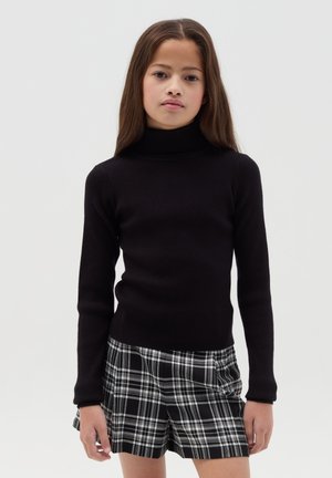 Suéter negro de cuello alto acanalado combinado con pantalones cortos de cuadros en blanco y negro. La parte superior presenta mangas largas y una silueta ajustada.