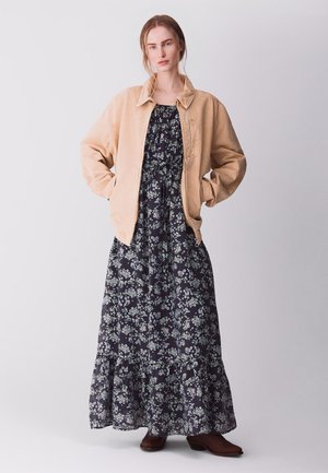 Donna con giacca beige sopra un lungo vestito nero a fiori e stivali marroni, in piedi con le mani nelle tasche della giacca su uno sfondo uniforme.
