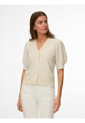 Femme portant un cardigan crème à manches courtes boutonné et un pantalon clair, debout devant un fond blanc, regardant sur le côté.