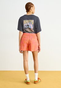 T-shirt graphique à manches courtes de couleur gris foncé, associé à un short rose à motifs. Le modèle porte des chaussettes blanches et des baskets colorées.