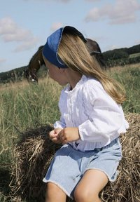 Bambino seduto su fieno, indossando una blusa bianca con volant e pantaloni di jeans corti con i bordi sfilacciati. Un cerchietto blu mette in risalto i lunghi capelli lisci.