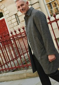 Palton gri cu model houndstooth și guler, având buzunare laterale și o croială ajustată, purtat peste un pulover alb și pantaloni negri. Balustradă roșie în fundal.