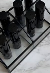 Sett med seks svarte, sylindriske kosmetikkflasker arrangert i en klar rektangulær skuff med speilbunn. Jevn tekstur, minimal merking.