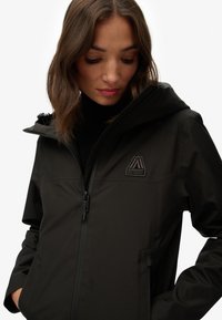 Black waterproof jacket with a zip closure, featuring a logo patch, high collar, and a smooth, matte texture. Hood is adjustable.