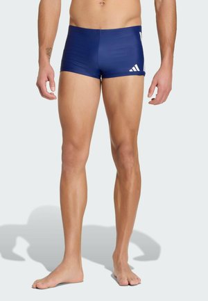 Mand iført marineblå Adidas badetrusser, stående barfodet mod en ensfarvet lys baggrund.