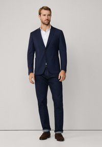 Niebieski blazer z gładkiego materiału z dwoma guzikami, w połączeniu z białą koszulką polo. Ciemnoniebieskie dżinsy z podwiniętymi mankietami i brązowe buty.