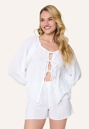 Lyshåret kvinde smilende, iført løs hvid bluse med tre bindebånd foran og matchende hvide shorts mod ensfarvet baggrund.