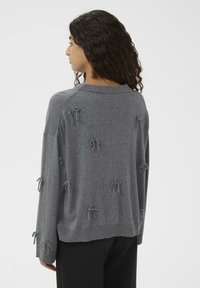 Ženska s kodrastimi lasmi, oblečena v ohlapen siv pulover z majhnimi zavezaniimi pentljami, razporejenimi po hrbtu in rokavih, v kombinaciji s črnimi hlačami.