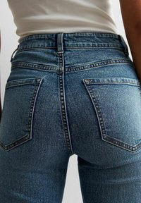 Höga, blå denimjeans med fem fickor, diskret slitningar och kontrasterande orange sömmar längs sömmarna och fickkanterna.