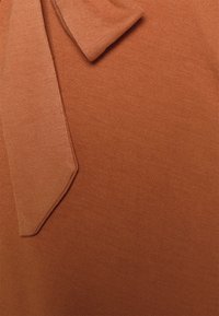 Tecido ribado castanho com um detalhe em nó na gola. A textura é suave, com um brilho subtil que realça a sua aparência.