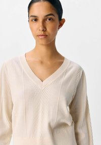 Cremfarbener Pullover mit V-Ausschnitt, der vertikale Rippmuster und eine weiche Textur aufweist. Die Ärmel sind lang und etwas lässig geschnitten.